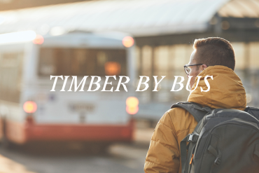 A man wearing a yellow coat looks at a bus. The words Timber by Bus are written across the image.