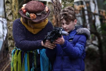 Two people are stood in front of trees looking at a crow puppet.