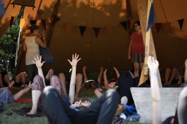 A group of people lie on the floor, inside a tent. They have their hands in the air. There is bunting in the background and a figure in a red t shirt and striped shorts stands in the background.