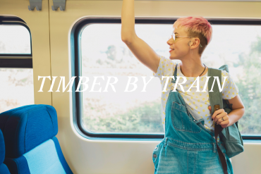 A person stands on a train and holds a rail. They are wearing denim dungarees.