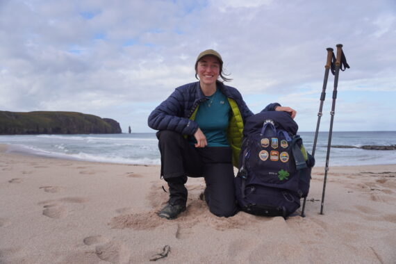 A person kneels on the sand holing a backpack to their side.