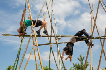 Two people are elevated leaning back over a bamboo structure. The sky is blue with a few clouds.