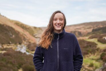 A woman stands against a mountainous landscape looking at the camera smiling.