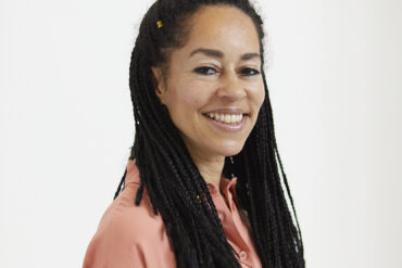 A woman stands side on looking at the camera.