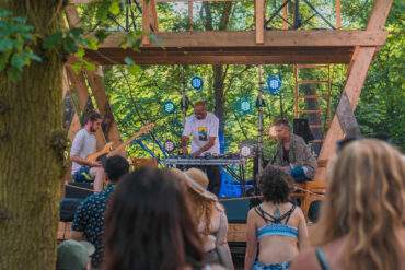 People are stood watching three people on stage outdoors. On the stage there is one person with a DJ deck, one person on the guitar and one person holding a clarinet.