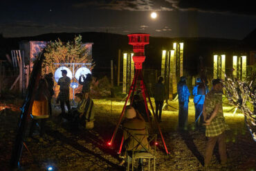 Multiple people stand around the shadows of illuminated structures under the moon