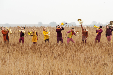 A group of brass musicians stand in a field of straw playing their instruments.