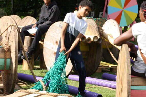 Two boys play with wooden structures and green rope. It's summer and they are in a woodland.