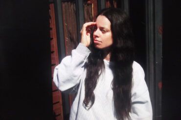 A person with long dark hair and fair skin looks to the left of the shot. They have their hand held up near their face. They wear a grey baggy jumper and have a serious face.