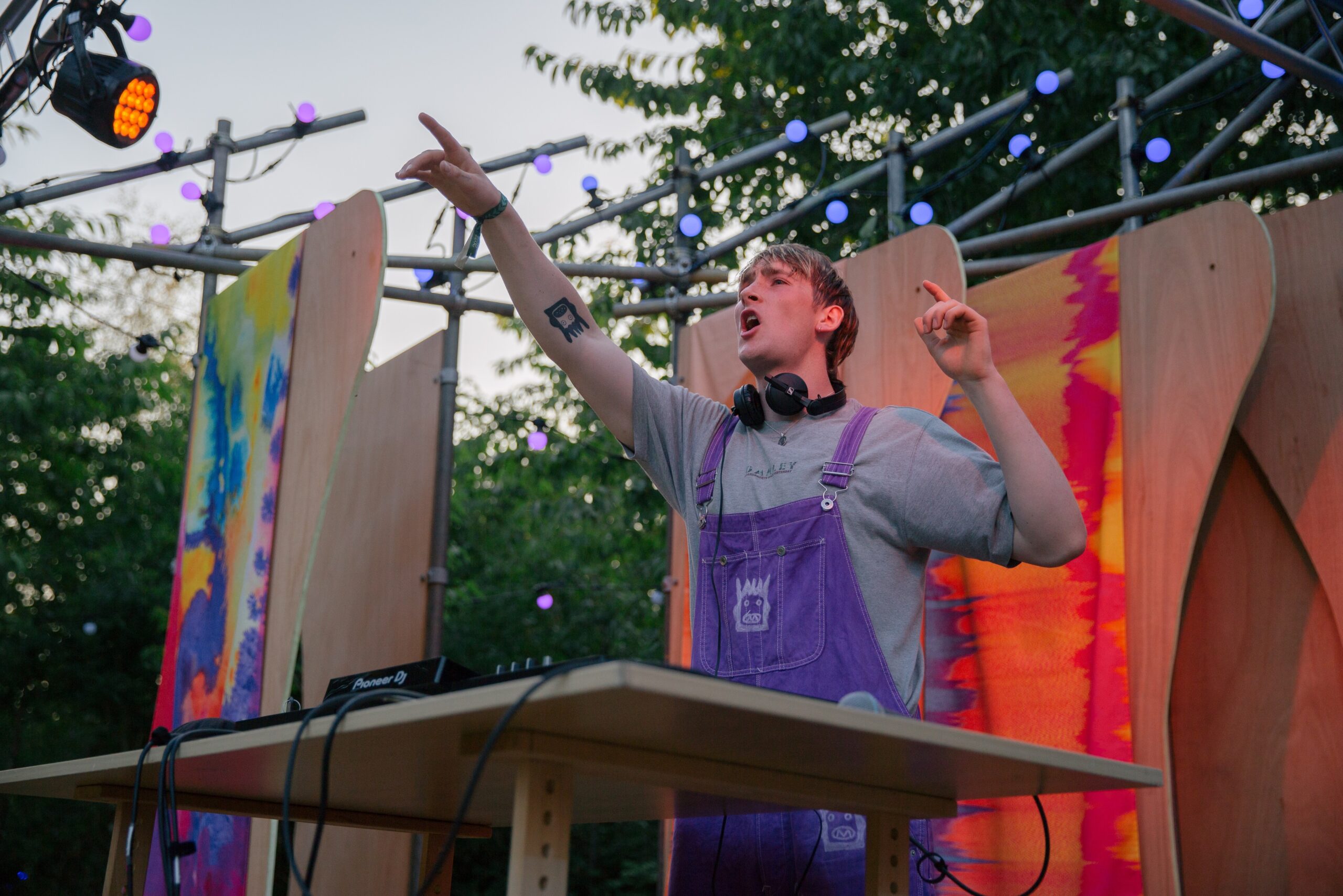 A person stands with a purple pair of dungarees. They are pointing to the crowd and standing behind a wooden desk.